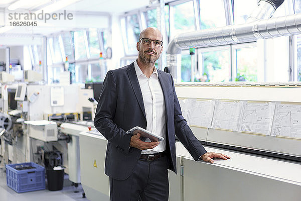 Selbstbewusster Geschäftsmann  der ein digitales Tablet in der Hand hält  während er vor Maschinen in einer beleuchteten Fabrik steht