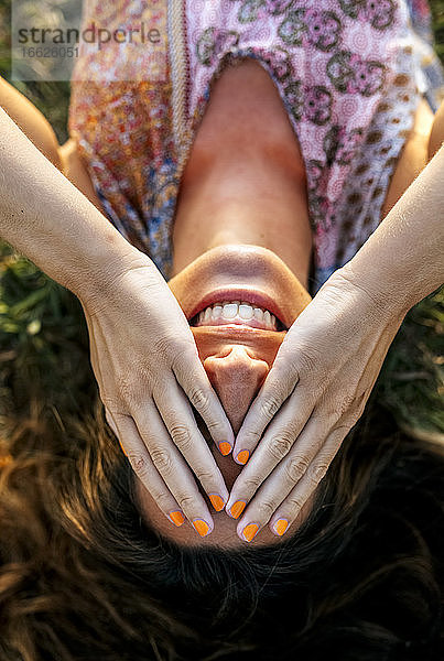 Lächelnde junge Frau  die ihre Augen mit den Händen bedeckt  während sie auf einem Feld liegt