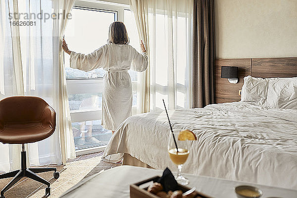 Ältere Frau im Ruhestand öffnet den Vorhang und blickt durch das Fenster eines luxuriösen Hotelzimmers
