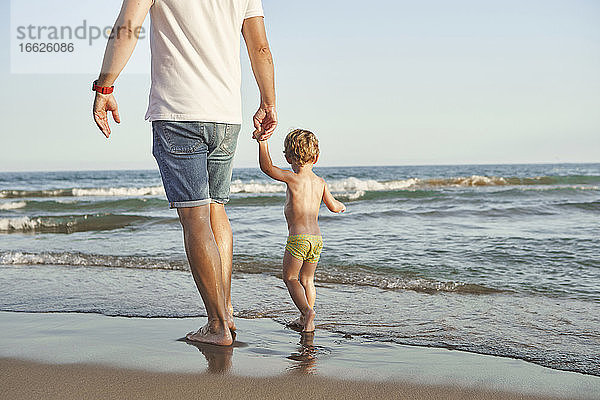 Vater und Sohn halten sich an den Händen  während sie am Strand spazieren gehen