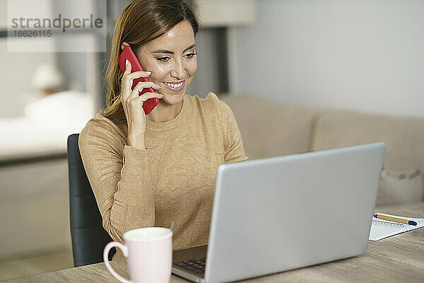 Lächelnde Geschäftsfrau  die am Telefon spricht  während sie zu Hause am Laptop arbeitet