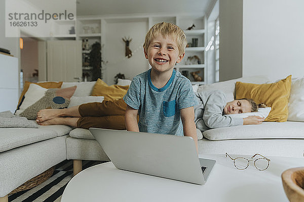 Lächelnder Junge mit Laptop und schlafender Mutter im Hintergrund