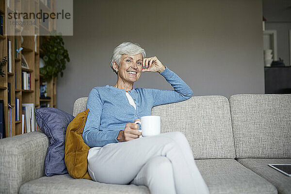 Lächelnde ältere Frau  die zu Hause auf dem Sofa sitzt und eine Kaffeetasse hält