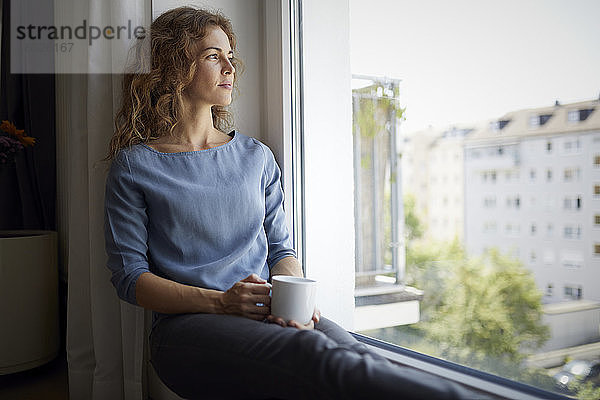 Frau mit Kaffeetasse  die durch das Fenster schaut  während sie auf der Fensterbank zu Hause sitzt