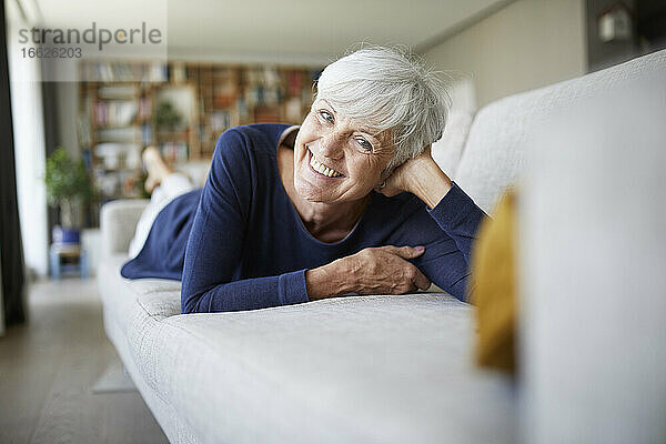 Lächelnde ältere Frau mit dem Kopf in der Hand auf dem Sofa zu Hause liegend