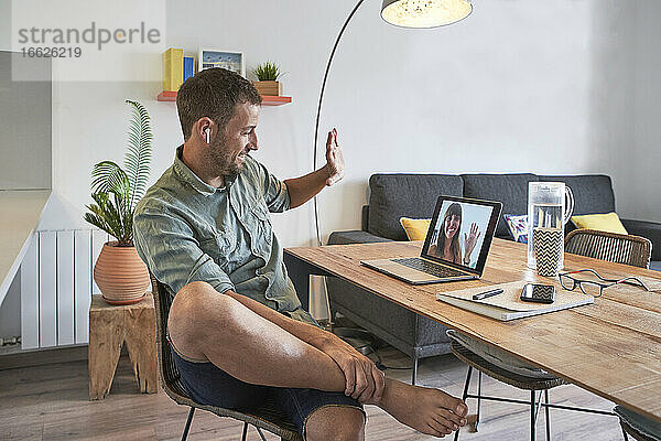 Lächelnder gut aussehender Geschäftsmann winkt einer Kollegin per Videoanruf auf dem Laptop zu  während er von zu Hause aus arbeitet
