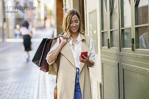 Frau mit Einkaufstasche  die ein Mobiltelefon benutzt  während sie auf einem Fußweg in der Stadt steht