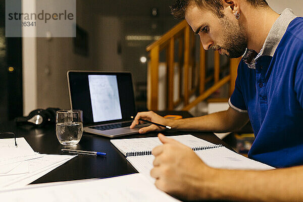 Junger Mann konzentriert sich auf seine Hausaufgaben  während er zu Hause am Laptop sitzt