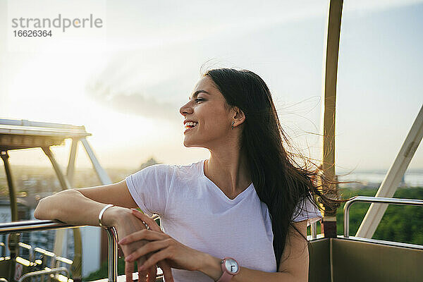 Lächelnde junge schöne Frau  die wegschaut  während sie das Riesenrad bei Sonnenuntergang genießt