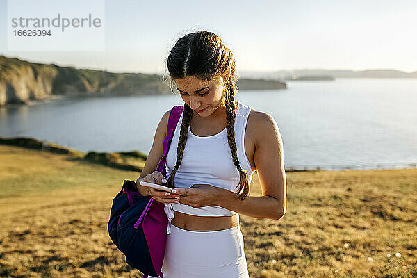 Junge Frau mit Rucksack  die ein Mobiltelefon benutzt  während sie bei Sonnenuntergang am Meer steht