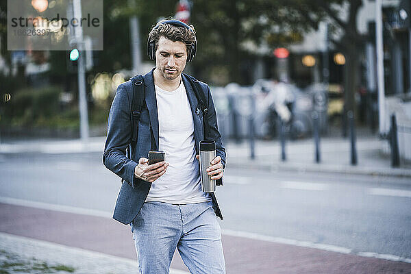 Mann mit Kopfhörer  der ein Mobiltelefon benutzt  während er in der Stadt auf der Straße geht
