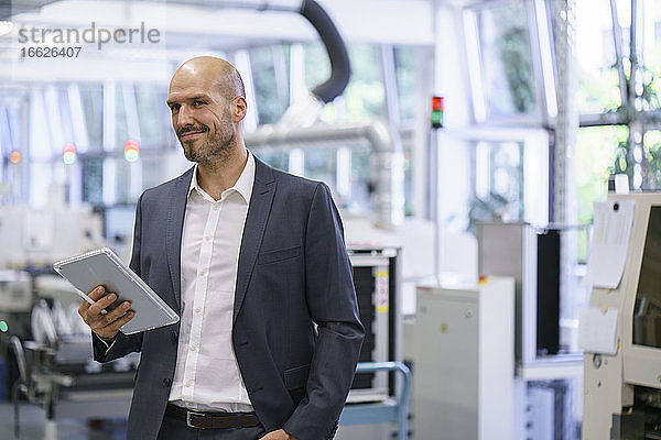 Lächelnder Geschäftsmann  der ein digitales Tablet hält und in eine helle Fabrik schaut