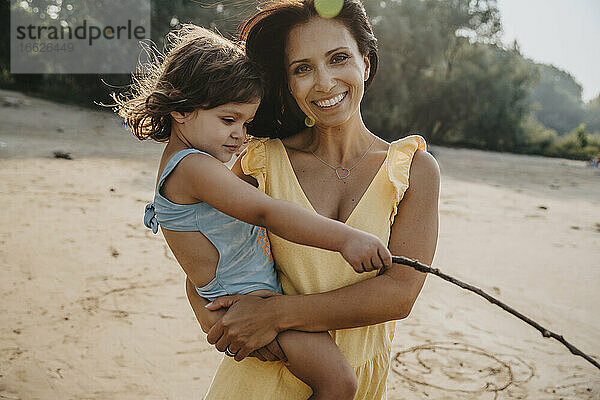 Lächelnde Mutter hält ihre Tochter in den Armen  während sie am Strand steht