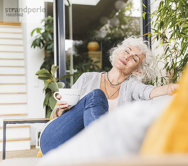 Reife Frau mit geschlossenen Augen mit Kaffeetasse sitzt auf der Couch zu Hause