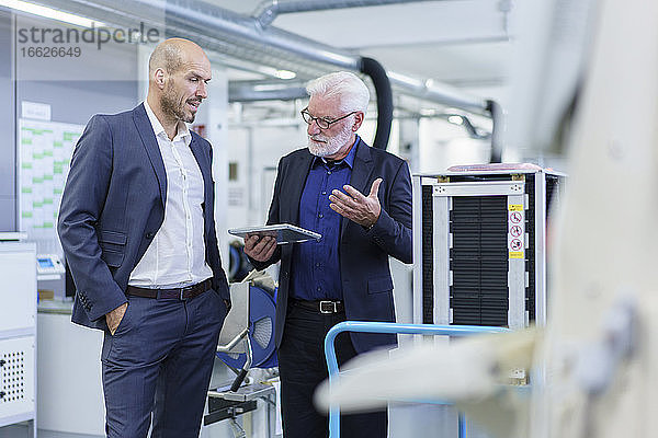 Männlicher leitender Angestellter  der mit einem Kollegen über ein digitales Tablet diskutiert  während er an einer Maschine in einer Fabrik steht