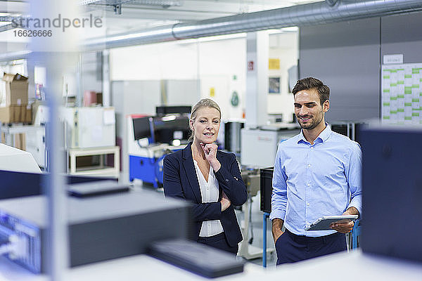 Lächelnde Berufskollegen diskutieren  während sie sich Maschinen in einer beleuchteten Fabrik ansehen