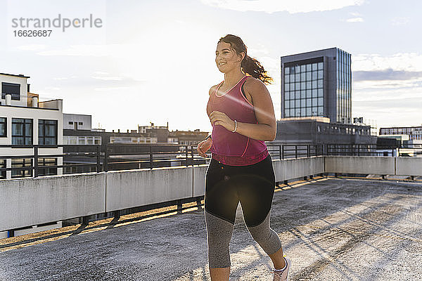 Lächelnde Sportlerin  die auf einer Terrasse gegen den Himmel in der Stadt läuft