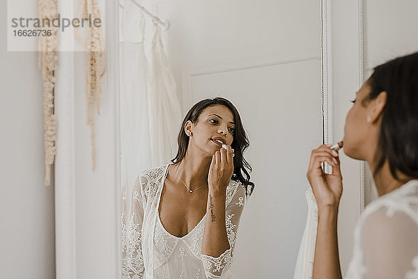 Junge Braut trägt Lippenstift auf  während sie sich zu Hause im Spiegel betrachtet