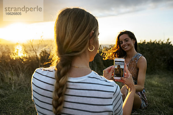 Junge Frau  die ihre Freundin bei Sonnenuntergang mit dem Smartphone fotografiert