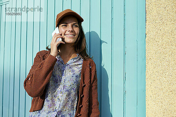 Junge Frau lächelt  während sie am Telefon gegen eine blaue Metalltür spricht