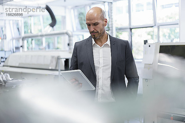 Ein glatzköpfiger Geschäftsmann schaut auf ein digitales Tablet  während er in einer hellen Fabrik steht