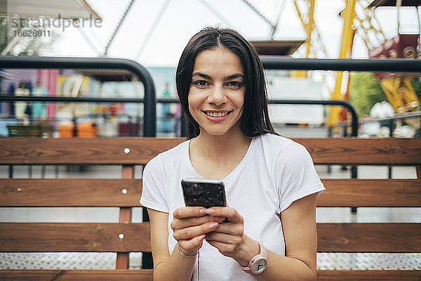 Schöne Frau mit Smartphone auf einer Bank im Vergnügungspark sitzend