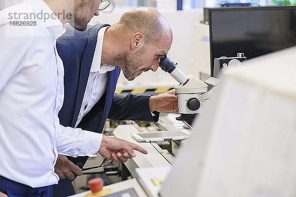Junger Techniker  der auf einen männlichen Kollegen zeigt  der durch ein Mikroskop im Labor schaut