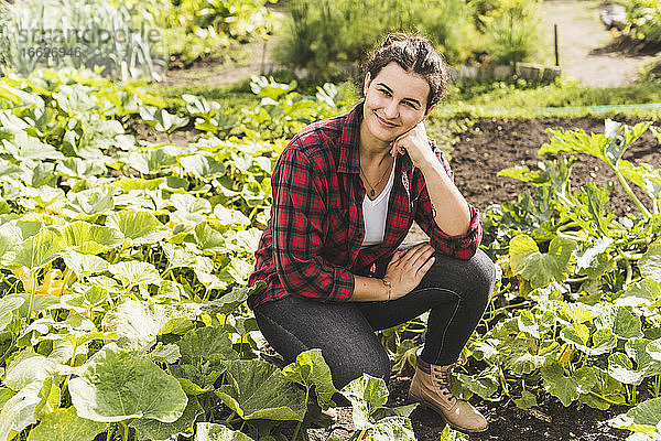Junge Frau mit Hand am Kinn kniend inmitten von Gemüse  das im Garten wächst