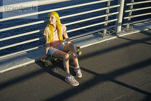 Junge Frau auf dem Skateboard sitzend gegen das Geländer an einem sonnigen Tag