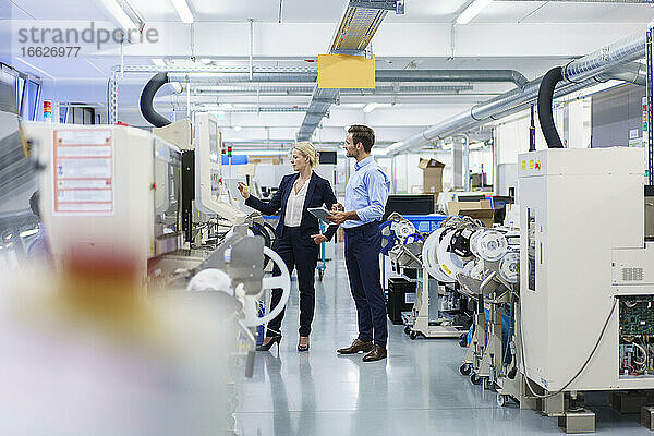 Reife Geschäftsfrau  die mit einem männlichen Ingenieur in einer beleuchteten Fabrik über Maschinen diskutiert