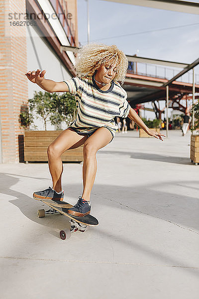 Blonde Frau mit ausgestreckten Armen beim Skateboardfahren auf dem Fußweg an einem sonnigen Tag