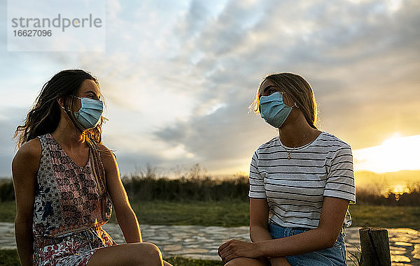 Frauen mit Gesichtsschutz  die während des Sonnenuntergangs gegen den Himmel sitzen und soziale Distanz wahren