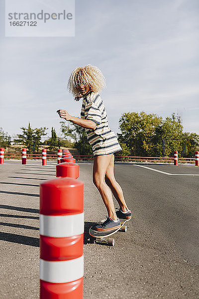 Blonde Skateboarderin  die einen Hindernisparcours auf der Straße gegen den Himmel durchfährt