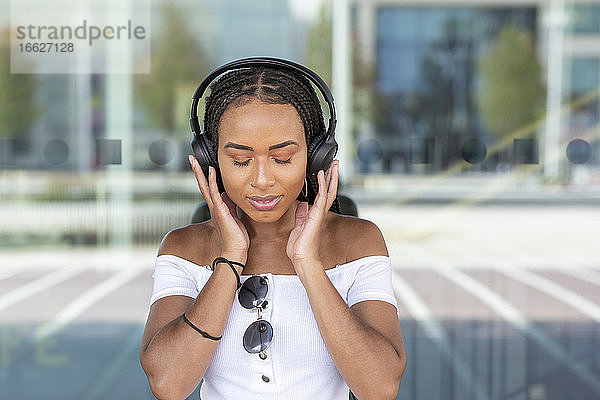 Junge Frau mit Kopfhörer hört Musik  während sie gegen eine Glaswand in der Stadt steht