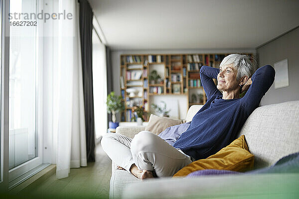 Ältere Frau mit Händen hinter dem Kopf mit gekreuzten Beinen auf dem Sofa zu Hause sitzen