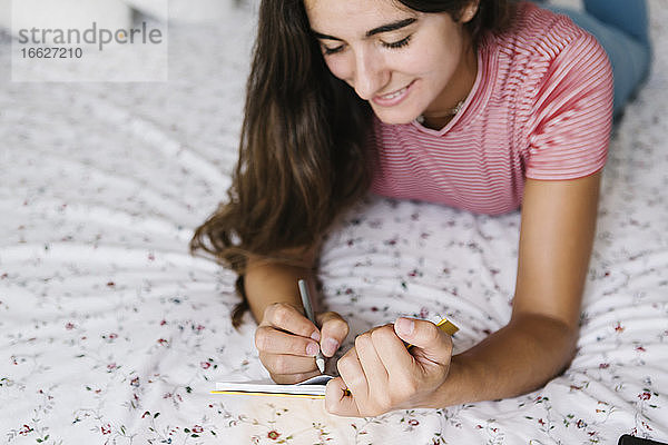 Junge Frau macht sich Notizen in einem Buch  während sie zu Hause auf dem Bett liegt