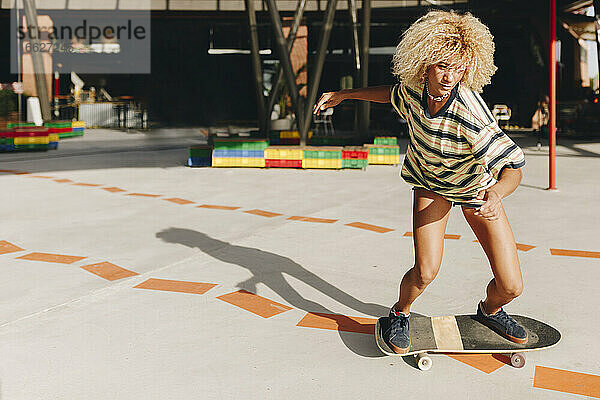 Selbstbewusste blonde Frau beim Skateboardfahren auf dem Fußweg an einem sonnigen Tag