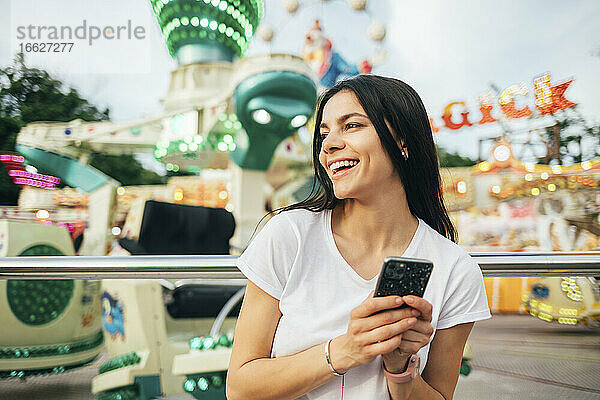 Fröhliche junge Frau mit Smartphone in der Hand  die in einem Vergnügungspark wegschaut