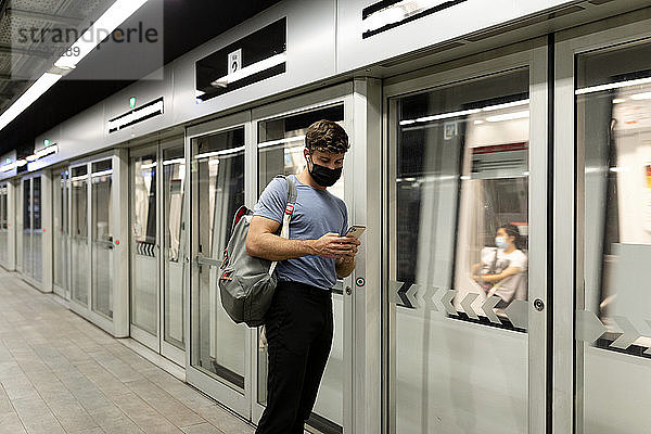 Junger männlicher Pendler  der sein Smartphone benutzt  während er in einem fahrenden Zug in einer U-Bahn-Station steht  während COVID-19