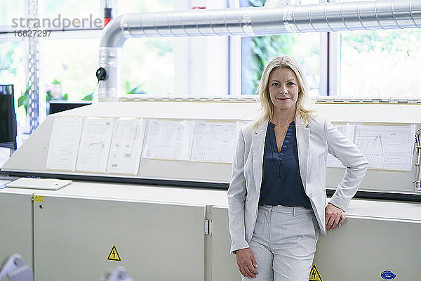 Selbstbewusste blonde Geschäftsfrau  die in einer beleuchteten Fabrik vor Maschinen steht