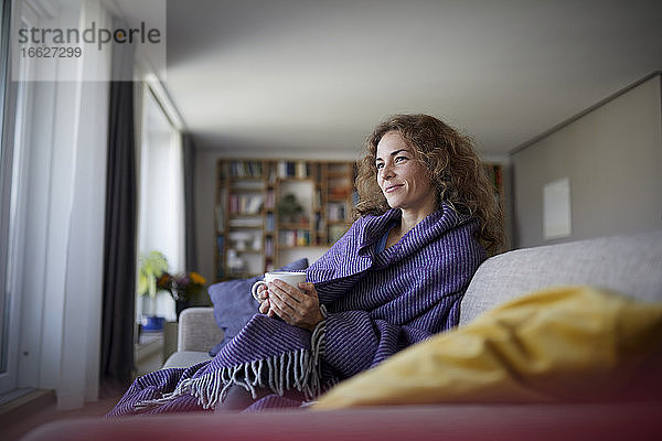 Lächelnde Frau mit Kopftuch  die zu Hause auf dem Sofa sitzt und Kaffee trinkt