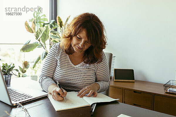 Frau  die in einem Boo schreibt  während sie einen Laptop auf einem Tisch zu Hause benutzt