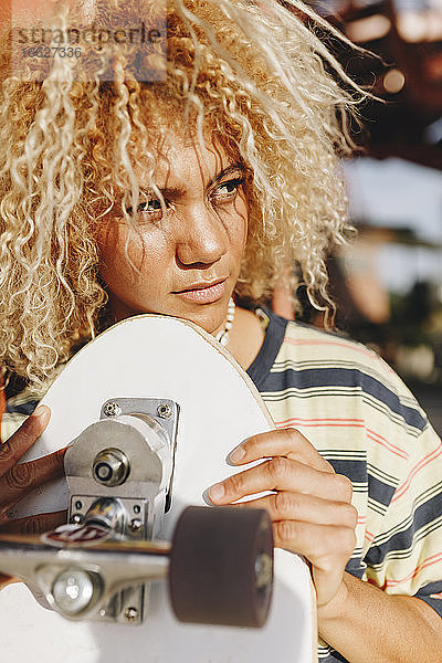 Hispanische Frau mit blonden lockigen Haaren  die wegschaut  während sie ein Skateboard an einem sonnigen Tag hält