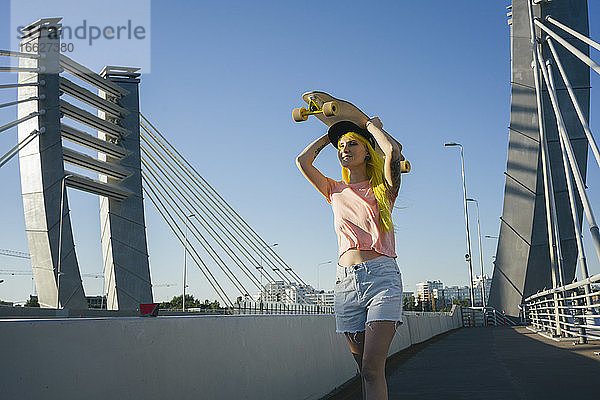 Junge Frau hebt Skateboard auf dem Kopf  während sie auf einer Brücke an einem sonnigen Tag steht
