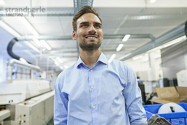 Lächelnder junger männlicher Ingenieur  der in einer beleuchteten Fabrik steht und wegschaut