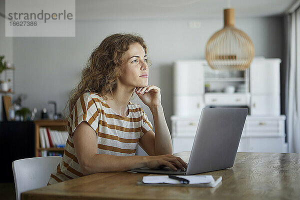 Nachdenkliche Frau mit Kopf in den Händen  die einen Laptop benutzt  während sie zu Hause am Tisch sitzt