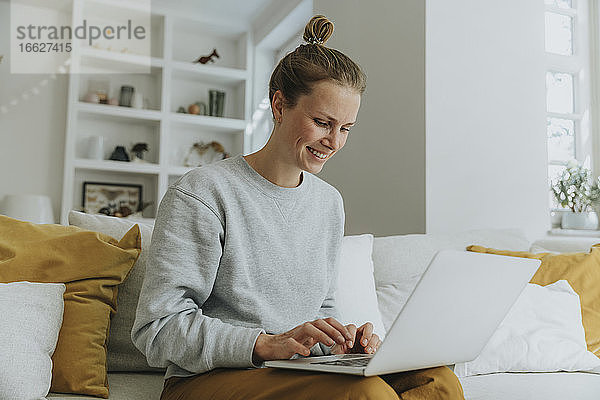 Lächelnde Frau  die einen Laptop benutzt  während sie zu Hause auf dem Sofa sitzt