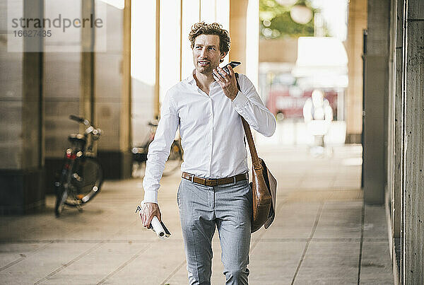 Lächelnder Mann  der am Telefon spricht  während er an einem Gebäude in der Stadt spazieren geht