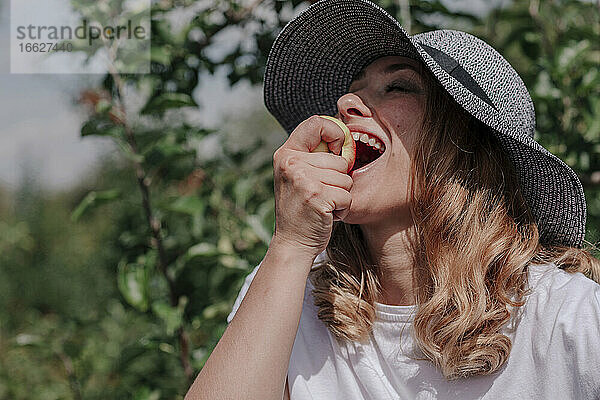 Nahaufnahme einer glücklichen Frau mit Hut  die einen Apfel im Obstgarten isst