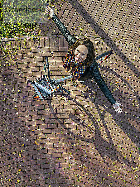 Luftaufnahme einer jungen Frau  die über einem Fahrrad steht und mit erhobenen Armen in die Kamera lächelt